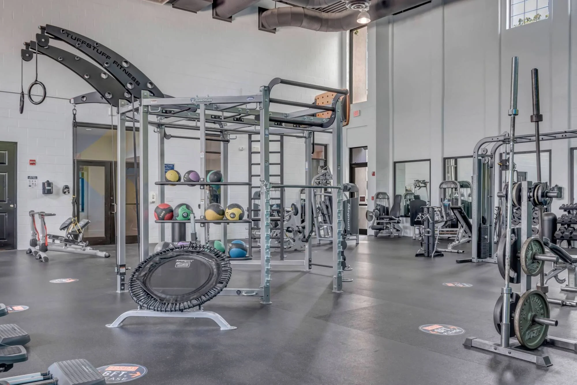 Spacious gym with workout equipment, medicine balls, weights, a trampoline, and high windows letting in light.