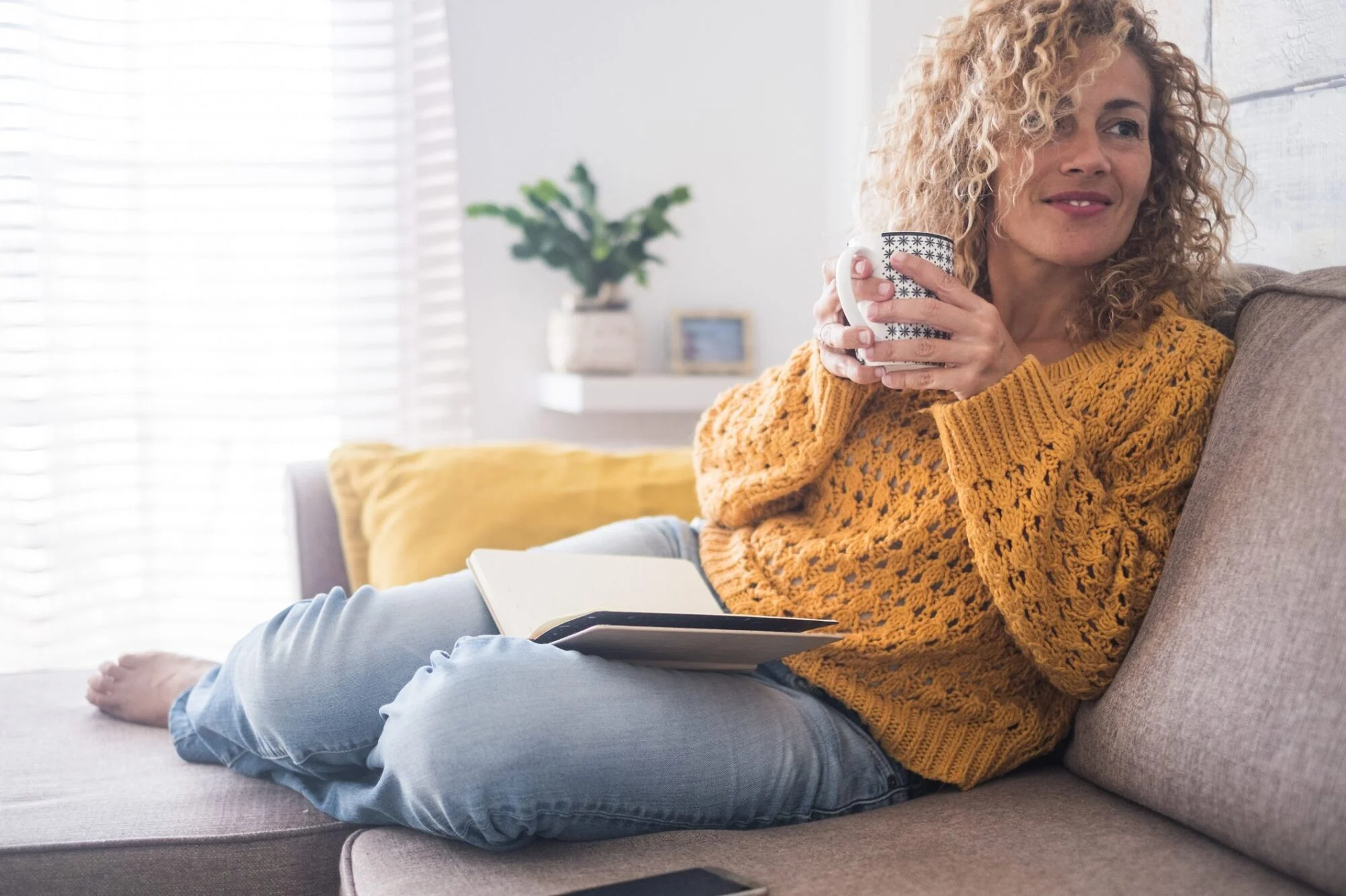 Woman in a yellow sweater sitting on a couch, holding a mug, with a notebook on her lap.